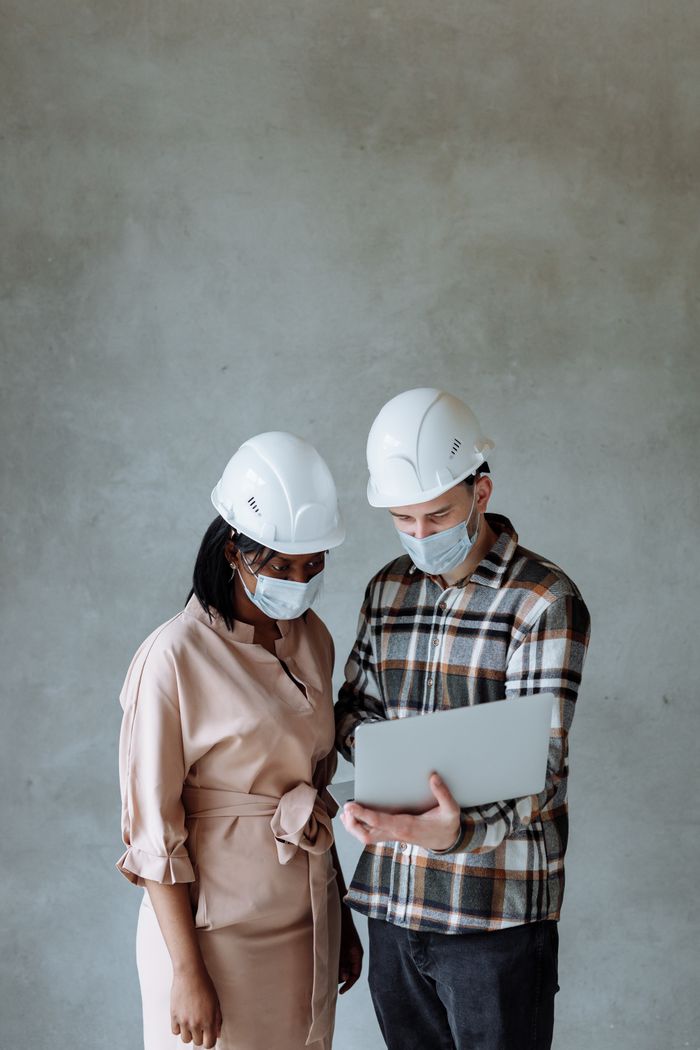 Two architects wearing face masks and hard hats collaborating on a laptop indoors.